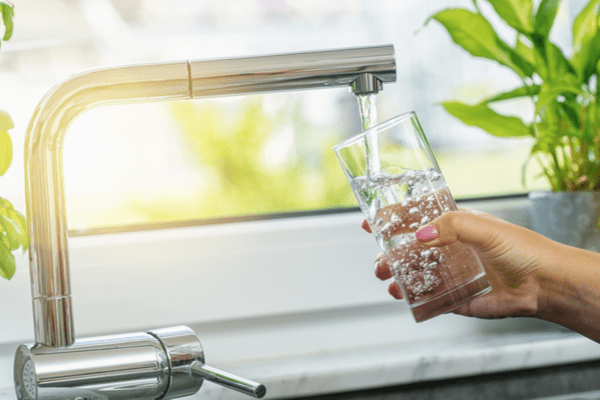 Clean water pours from a kitchen faucet and into a glass with the sun streaming in behind.