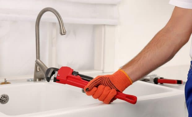 A man with a pipe wrench is near the kitchen sink, installing a water tap as part of plumbing upgrades according to Texas plumbing code.
