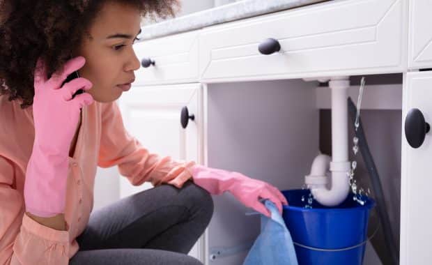 A woman experiencing plumbing emergencies, wearing pink gloves, is calling a plumber while standing in front of a leaking sink pipe.