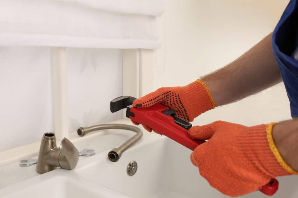 Man with pipe wrench near kitchen sink, installing water tap as part of plumbing upgrades.